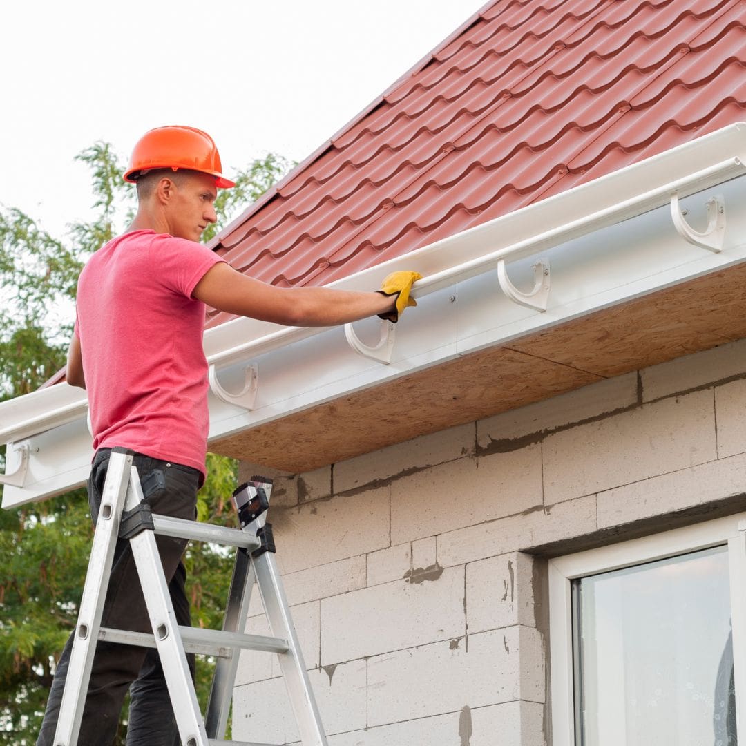 Installer in a hard hat on a ladder fitting a new white gutter to a roof edge